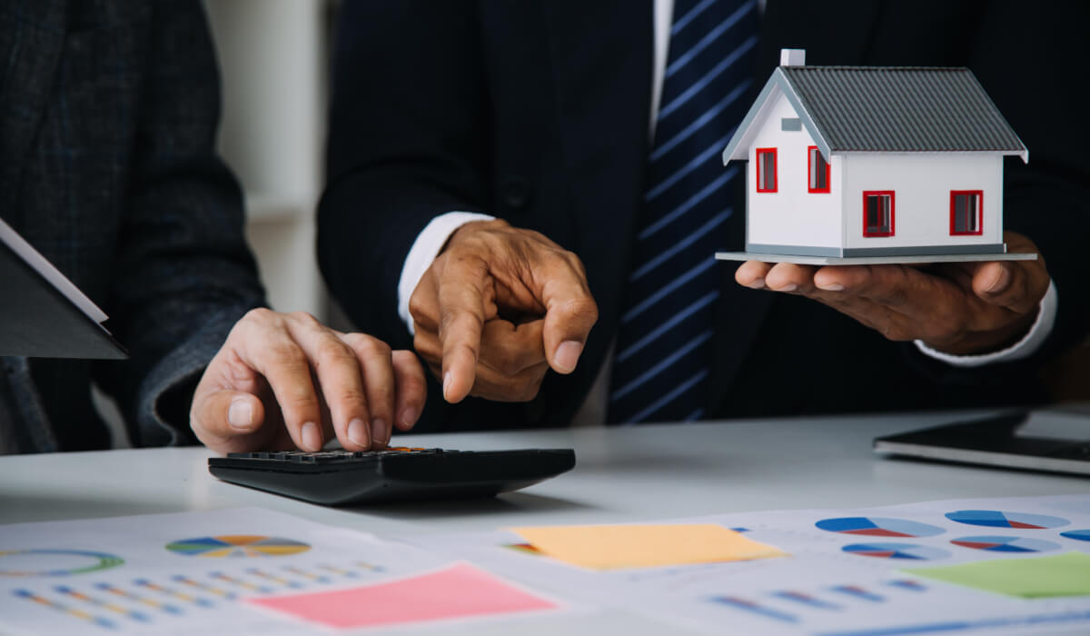 Two professionals in suits, one holding a small model of a house and the other using a calculator, with only their hands visible in a professional setting.