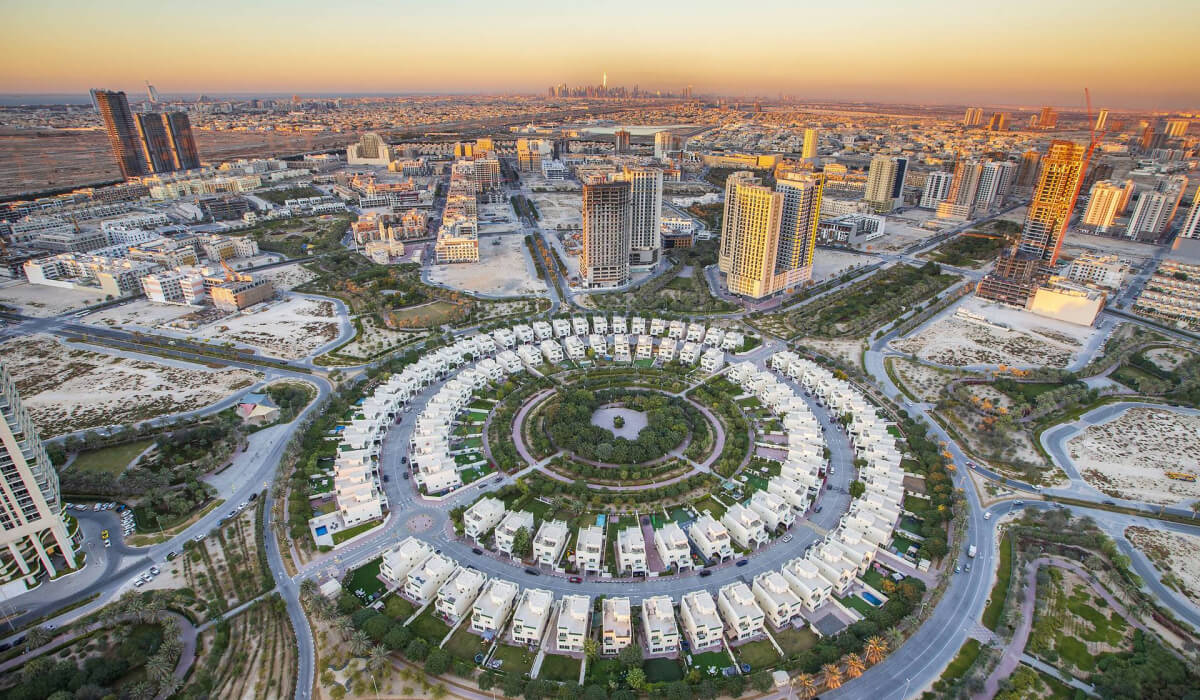 Top view of Jumeirah Village Circle, showcasing its symmetrical architecture, modern high-rise buildings, and wide streets, surrounded by a desert landscape that has transformed into green spaces.