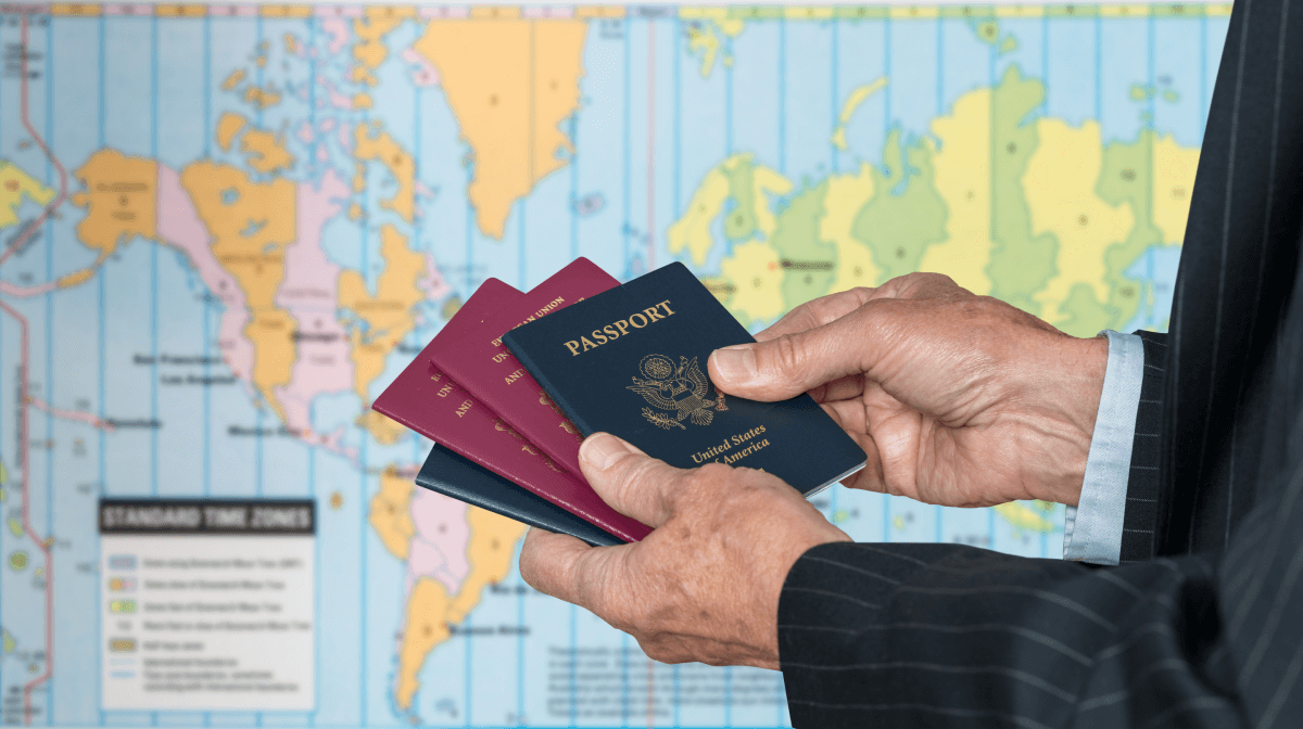 A professional man in a suit standing in front of a world map, holding four passports in his hand.