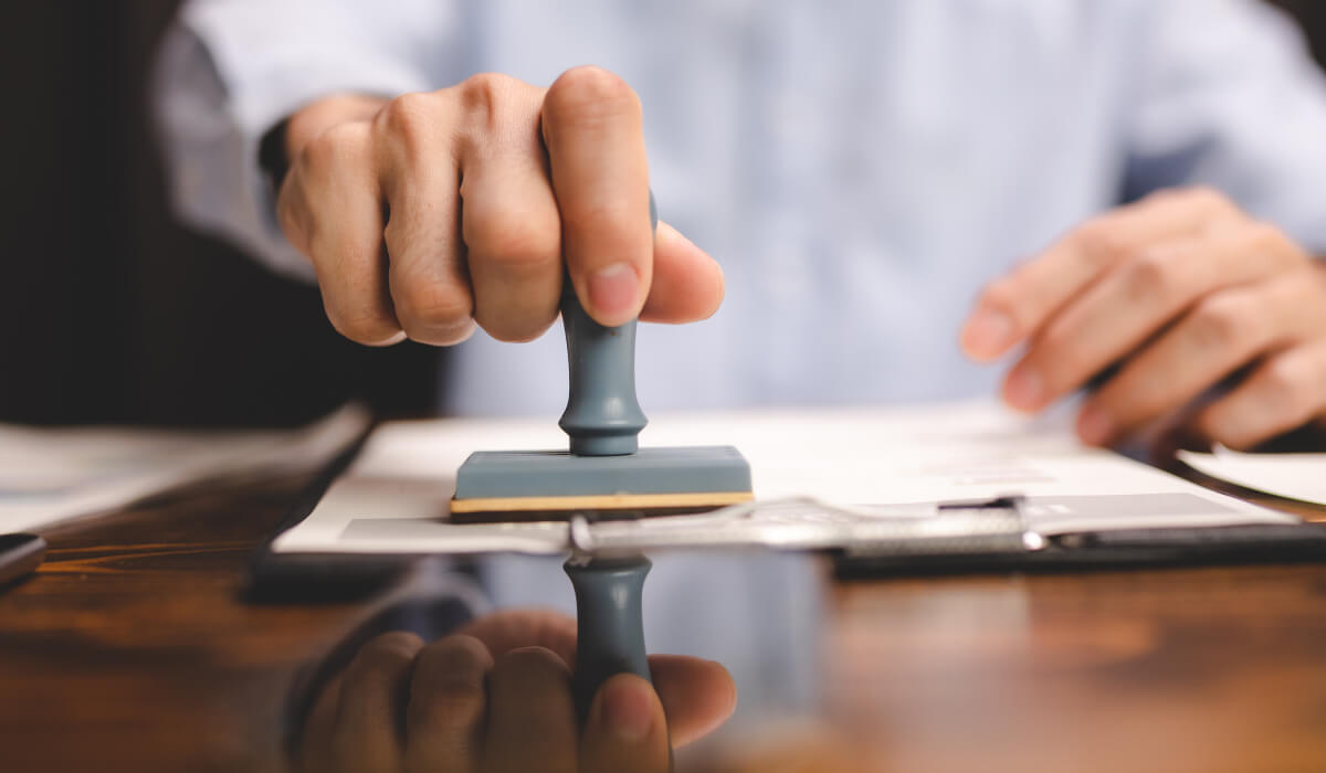 A hand presses a stamp onto a document on a wooden table, with the silhouette of the stamp clearly visible on the table's surface.