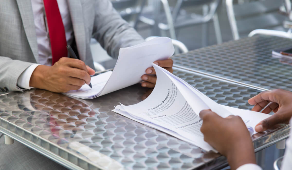 Two professionals are seated outdoors at a table, one signing a document while the other reads, with their hands and paperwork clearly visible.