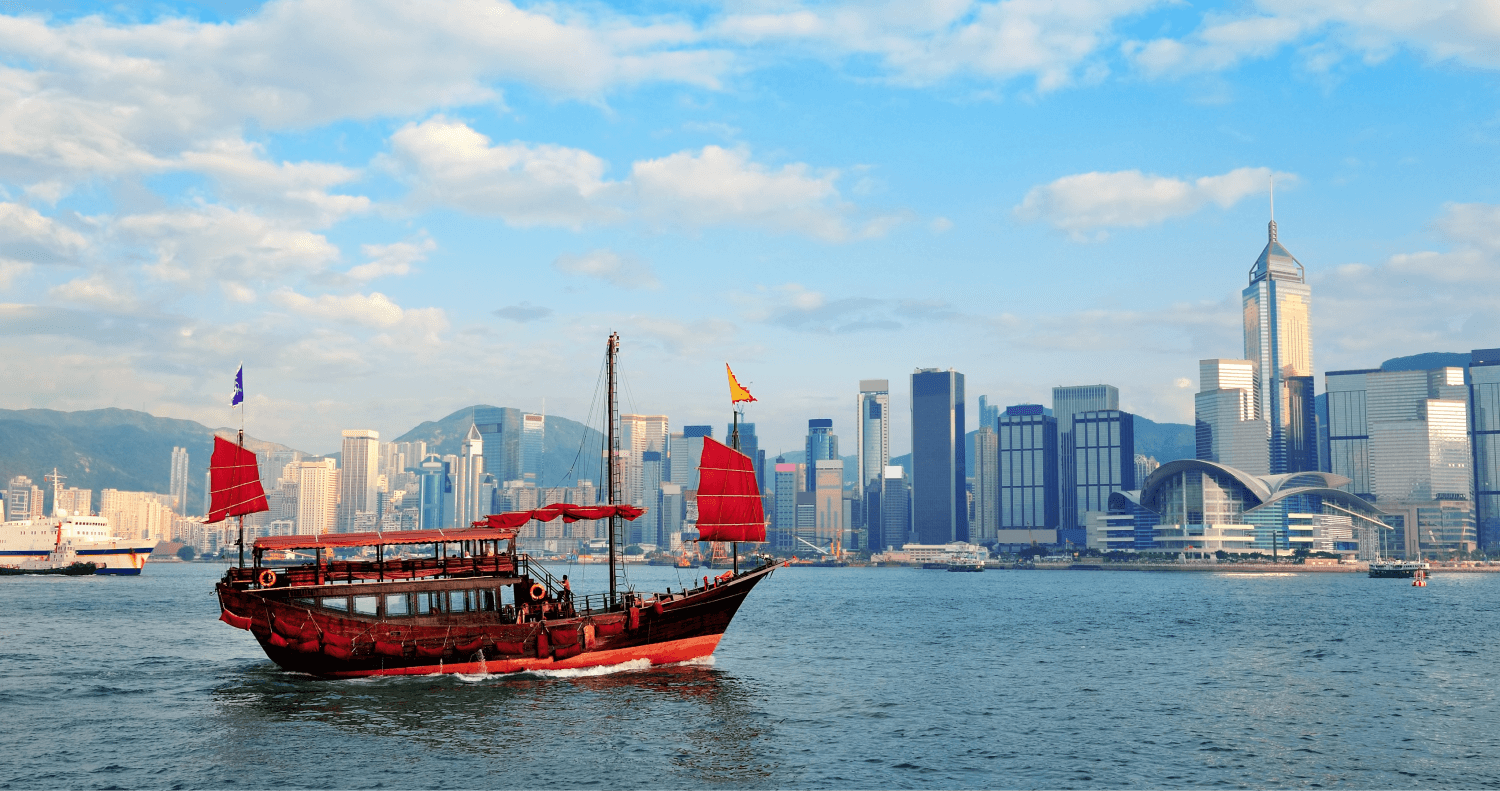 A traditional red Chinese-style boat floats gracefully on the water near Hong Kong, with the city's modern buildings, green spaces, and blue sky providing a stunning backdrop.