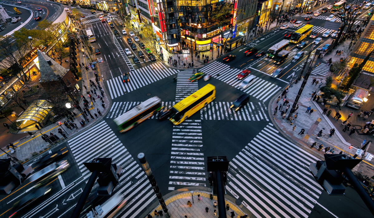 Top view of a busy Tokyo intersection with four crossing streets, blurred vehicles including yellow and green buses, cars, and buildings lining the sides.