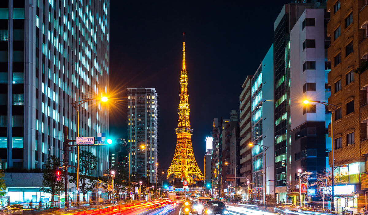 Nighttime Tokyo street with bright lights, towering buildings on both sides, and a shining tower in the center.