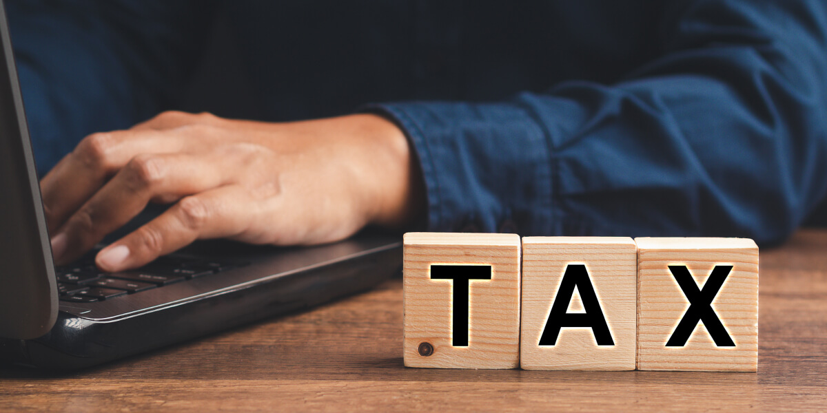 A work desk featuring a computer, a hand resting on it, and cubes with the word 'TAX' displayed on the table.