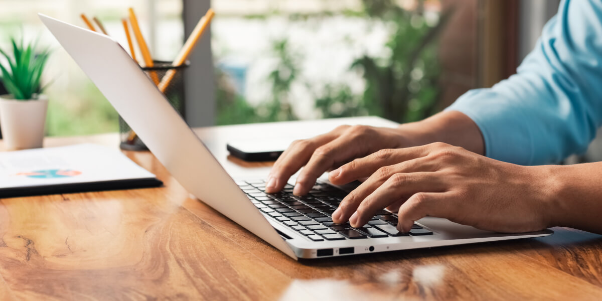 Hands typing on a computer placed on a table, surrounded by documents, with natural light streaming in from a nearby window.