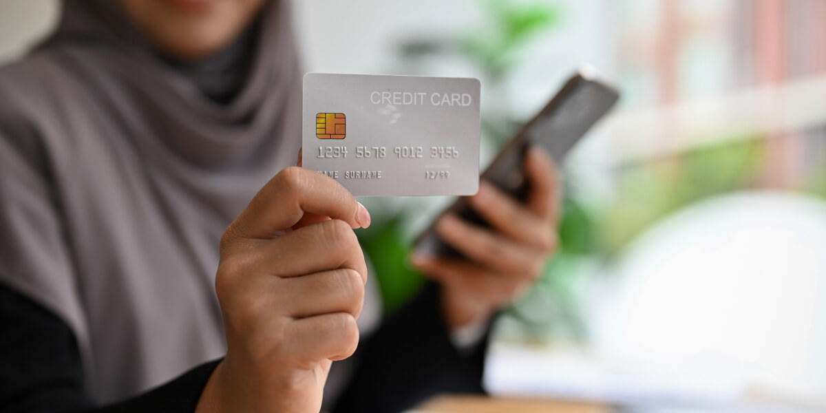 A Muslim woman dressed in gray holds a zoomed-in credit card, while her image is blurred; in her other hand, a phone is also blurred, creating a focus on the card.