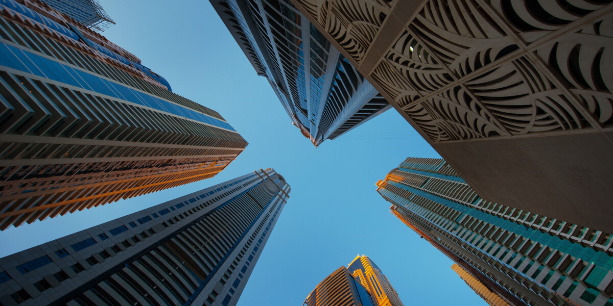 A low-angle photograph of tall modern skyscrapers set against a clear blue sky, capturing the imposing architecture and vertical lines of the buildings as they reach upwards.