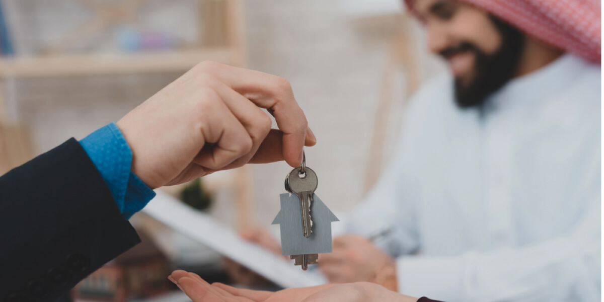 A close-up of keys hanging from a keyring shaped like a house, with a person’s hand holding them.