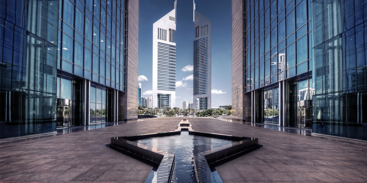 A modern corporate building with reflective glass windows, set against the backdrop of two skyscrapers in a sunny cityscape.