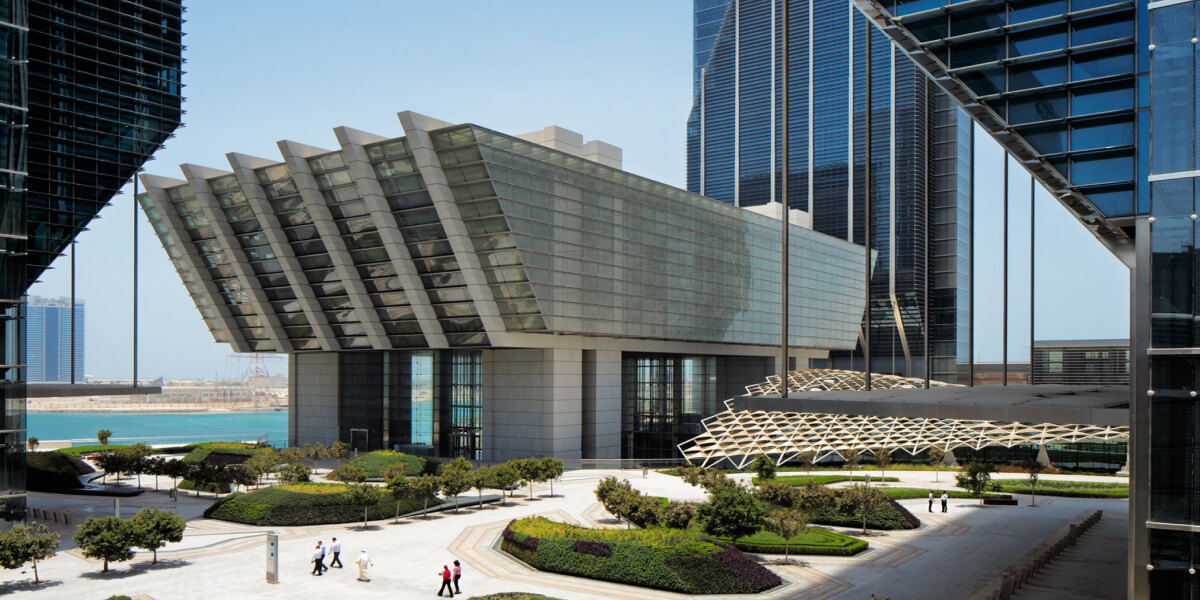 An angular, futuristic building with glass facades, positioned near a waterfront with a modern city skyline in the background.