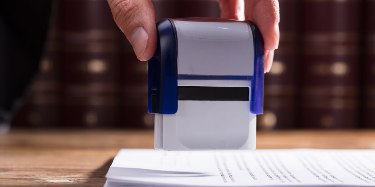 Close-up of a hand pressing a blue and white stamp in an office setting.