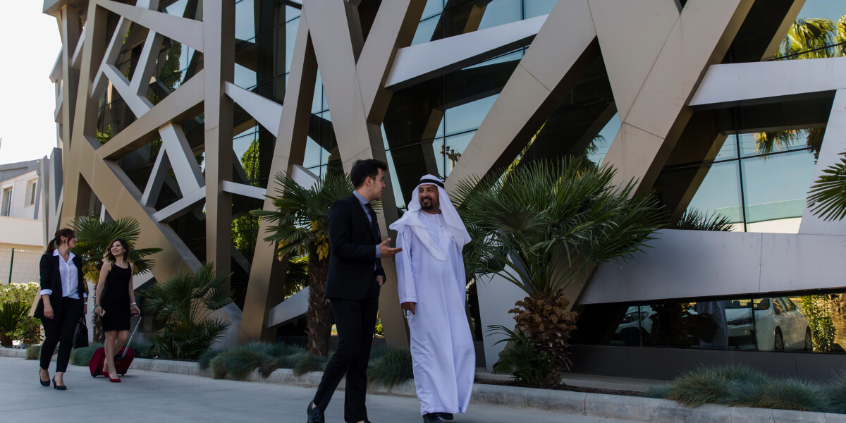 A group of business professionals, including a man in traditional Emirati attire, walking and conversing outside a modern building lined with palm trees.