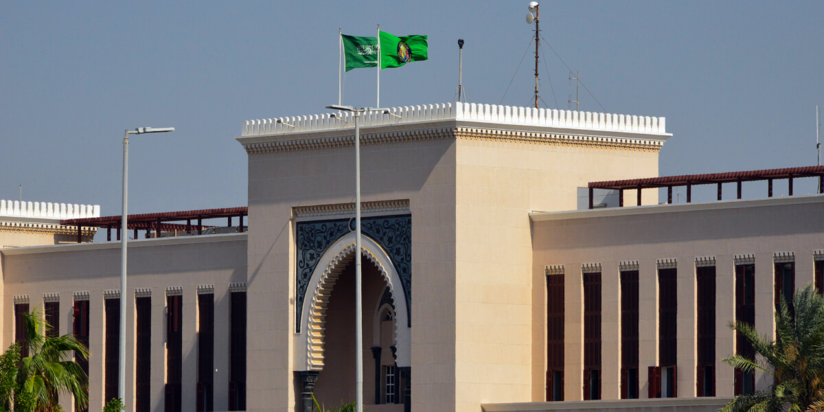 Two green flags, including the Saudi Arabian national flag, flying atop a traditional government-style building.