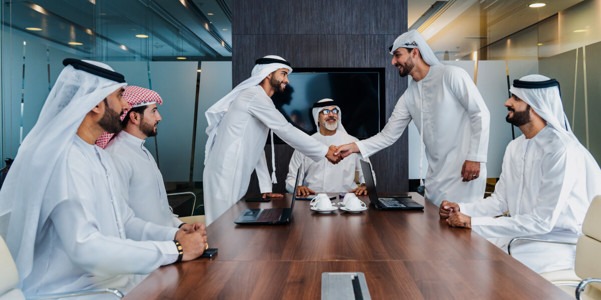 Gulf businessmen in traditional attire shaking hands across a meeting table, signifying a successful deal.