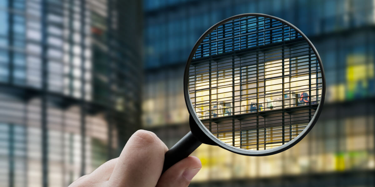 Close-up of a hand holding a magnifying glass over a modern office building, zooming in on an illuminated interior workspace.