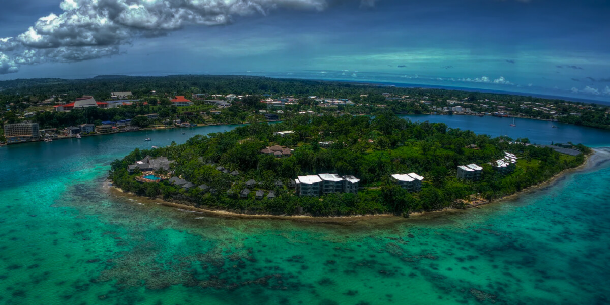 Aerial view of a tropical city in Vanuatu with dense greenery and turquoise waters under dramatic clouds.