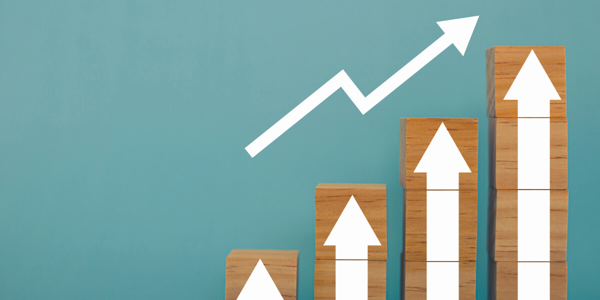 Wooden blocks stacked to show business growth, with white upward arrows and a trend line on a teal background.